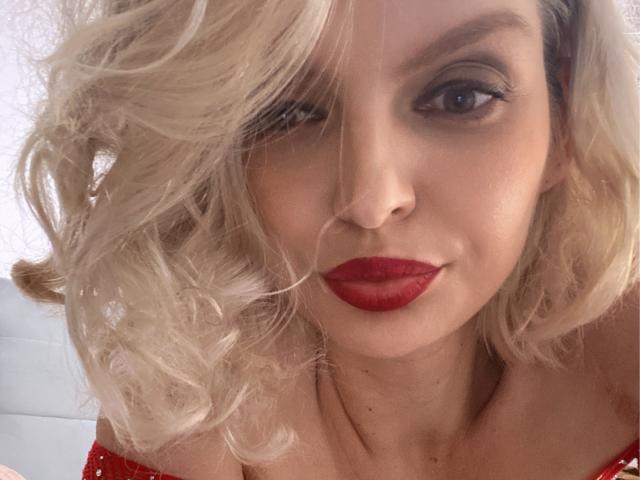 DouceAmelia has blonde, curly hair, red lipstick, and is wearing a red, off-shoulder top, showing a bit of cleavage, with a subtle smile. Her eye makeup is dark.