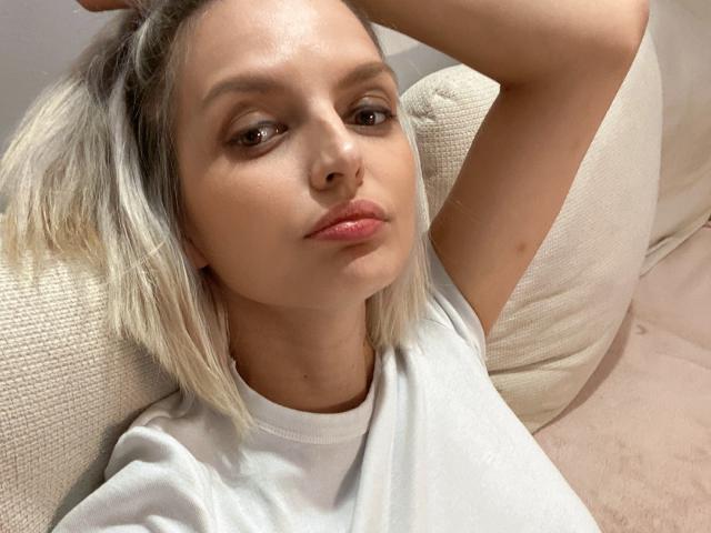DouceAmelia with blonde hair, white shirt, and pouty pink lips, arms raised, sitting on a beige couch, looking slightly to the side, with a soft, relaxed expression.