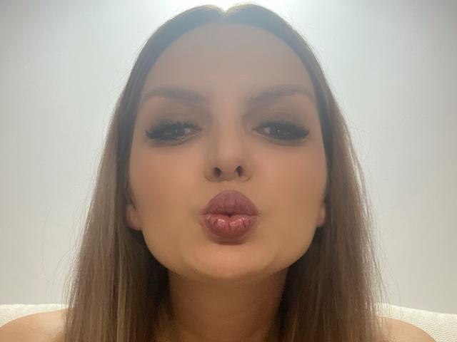 DouceAmelia has pouty pink lips, full eyebrows, long brown hair, and heavy black eyeliner, making a puffy kiss face. She's centered in the shot with a neutral background.