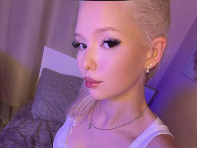 Blonde WillodeanAlmonte with long eyelashes, wearing white lace top, small nose ring, and necklace, looking at camera with slightly parted glossy pink lips, purple lighting.