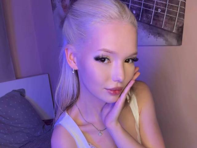 Blonde WillodeanAlmonte with long hair, wearing a white top, has her hand on her cheek, showing off her tits with big eyelashes and a slight smile.