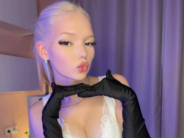 WillodeanAlmonte with blonde hair, black satin gloves, white lace bra, pouty lips, and cleavage, looking seductive, wearing a silver cross earring.