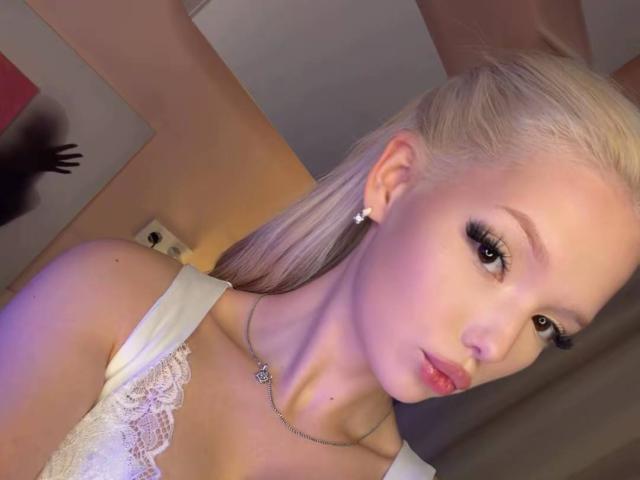 WillodeanAlmonte has blonde hair, large eyes with heavy eyeliner, pink lips, white lace top, necklace, and earrings, showing a serious expression in a well-lit room.