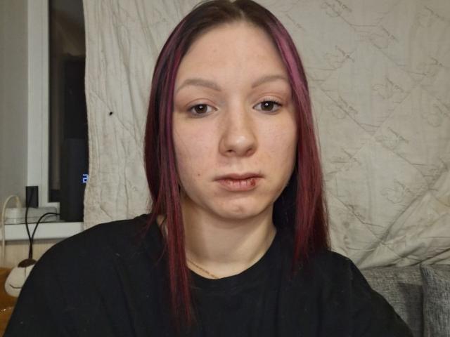 ChasidyBarranca with pale skin, straight red-purple hair, brown eyes, slight pout, black shirt, freckles, and a small lip piercing, sitting in a dimly lit room.