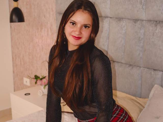 BeccaSimons has long brown hair, red lipstick, and a black sheer top, showing small tits and a red plaid skirt, sitting on a bed with a cute smile.