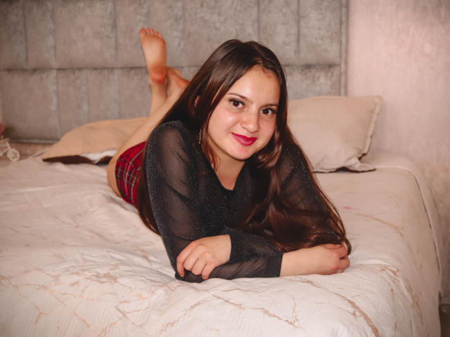 BeccaSimons is lying on a bed, her ass up, wearing a black sheer top and red plaid shorts, with her feet in the air, smiling at the camera.