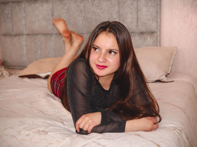 BeccaSimons with long brown hair, black sheer top, red plaid shorts, lying on bed, legs up, arms folded, smiling, pink lipstick, bare feet, gray headboard, white pillows.