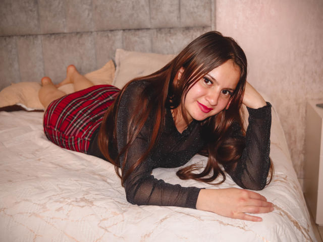 BeccaSimons with long brown hair, black sheer top, and red plaid skirt, lying on a bed, smiling, her ass raised, hand on head, legs up, on white quilted bed.