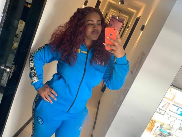 GabbyBonnye with curly red hair, wearing a blue track suit, holding an orange phone, taking a mirror selfie, showing a hand on her hip, standing confidently.