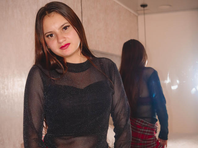 BeccaSimons with long brown hair, red lipstick, wearing a sheer black top, standing in front of a mirror, showing her back and plaid skirt, reflection visible.