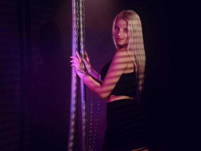AliceWhitey with long blonde hair, black crop top, and black skirt, holding a string of beads, looking over her shoulder, purple and pink lighting, showing her ass.