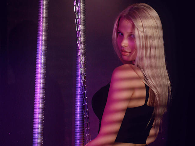 AliceWhitey with long blonde hair, black tank top, looking over shoulder, standing next to a chain with purple and pink striped light reflections on her skin and hair.