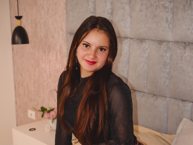 BeccaSimons with long brown hair, wearing a sheer black top, red lipstick, and earrings, sitting on a bed, smiling at the camera. Her ass is partially visible.