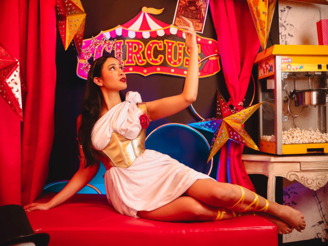 JennTylor with long dark hair, wearing a white toga-like dress with gold accents, sitting on a red bench, holding circus tent, red curtains, popcorn machine, yellow star decorations.