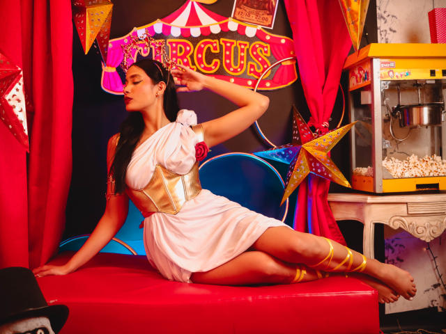 JennTylor in a white toga-style dress with gold accents, lying on a red bench, arm raised, showing off her tits, with popcorn machine and star decorations in background.