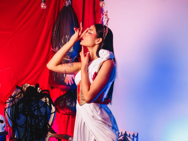 JennTylor with long black hair, eyes closed, wearing a white dress, touching her face, red and blue lighting, white scarf, black mask, and jewelry on a wooden table.