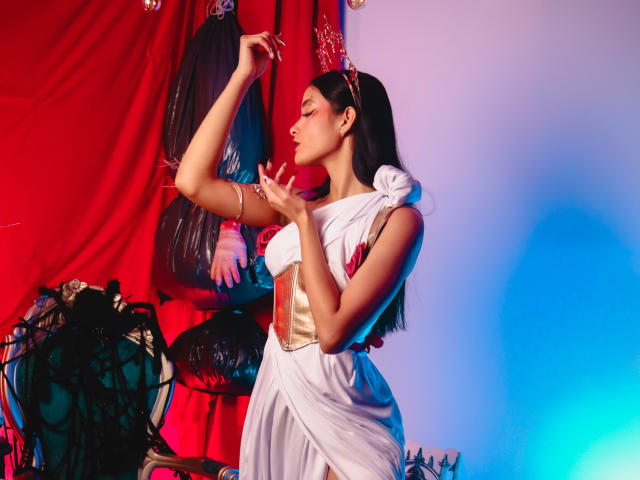 JennTylor in a white toga-style dress, gold belt, and headband, arms raised, eyes closed, touching her nose, standing next to a red curtain with black, shiny, mask-like objects.