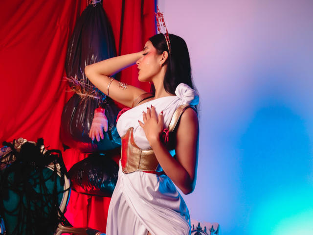 JennTylor with tan skin, dark hair, and medium tits, wearing a white dress with a gold belt, hand on forehead, standing next to a red curtain and blue light.