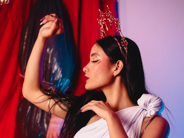 JennTylor with long black hair, wearing a white dress and gold headpiece, raises arm, showing off glittery eye makeup and pink blush, red curtain in background.