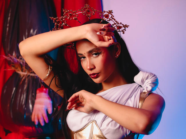 JennTylor with long black hair, wearing a gold crown and white dress, covering her tits with her arm, red lipstick, and gold arm bracelet, against a colorful background.