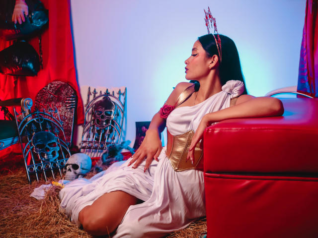 JennTylor with dark hair, wearing a white toga dress, gold belt, and red headpiece, sitting on straw, arms resting on red leather couch, surrounded by skulls and decorative pieces.
