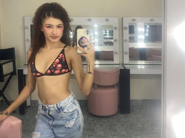 NathachaEvans with curly brown hair, small tits in a pink floral bikini top, and blue distressed jeans, taking a mirror selfie in a brightly lit room.