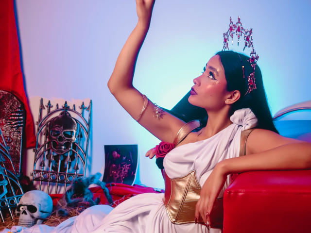 JennTylor with long black hair, wearing a white toga, gold arm bracelet, and a jeweled crown, raises her arm, showing off her tits, with a skull and red furniture.