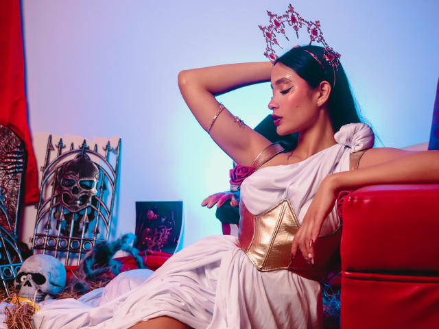 JennTylor with long black hair, wearing a white toga dress, gold corset, and red flower crown, holding a gold clutch, leaning on a red sofa, with a skull nearby.