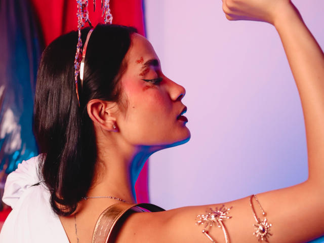 JennTylor with dark hair, raising arm, wearing a metallic headpiece and armlet with flowers, has red makeup on cheek, white robe, and pinkish-blue light. Her tits are visible.
