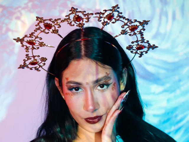 JennTylor with dark hair, wearing a fancy red and black headpiece, looking at the camera with dark lipstick, glitter on cheeks, and long nails touching her face.