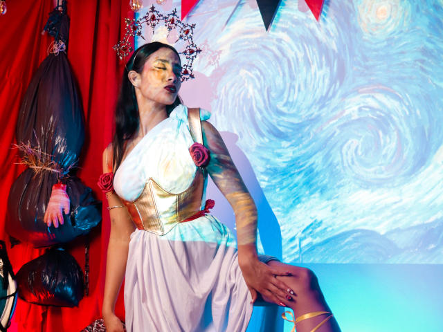 JennTylor in a white and gold dress with red roses, gold corset, and crown, standing with hand on thigh, red curtains, black bags, swirling blue and white background.