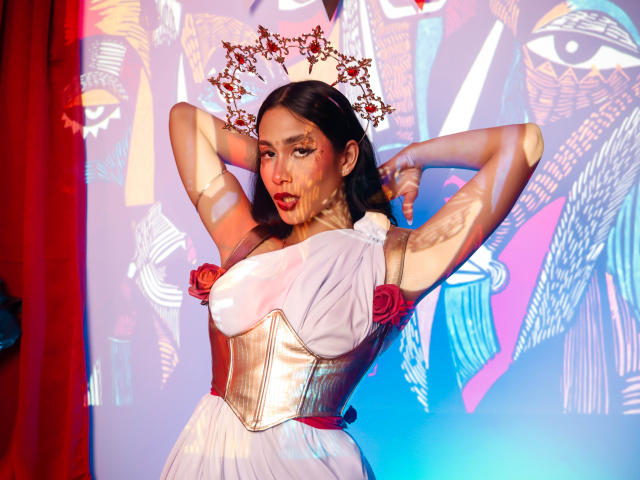 JennTylor with arms up, wearing a white dress, gold corset, red roses, and a floral crown, showcasing her tits and red lipstick, against a colorful, patterned backdrop.