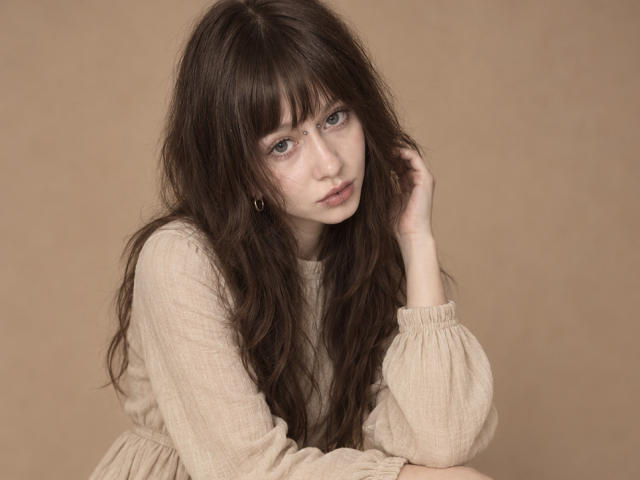 LorineThomeczek has long, messy brown hair, blue eyes, and a serious expression. She's wearing a beige, long-sleeved, ribbed shirt, with her hand touching her head.