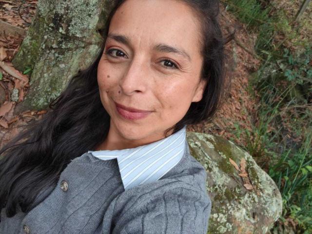 StangelarRivera with medium brown skin, black wavy hair, and a small mole on her right cheek, wearing a grey cardigan over a blue-striped shirt, smiling softly.