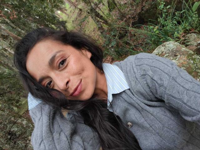 StangelarRivera has long black hair, brown eyes, and a slight smile. She's wearing a gray cardigan with a blue striped shirt, and has a cute, relaxed look.