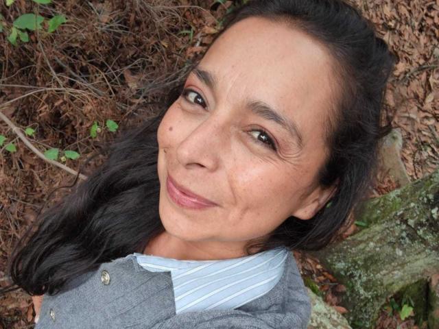 StangelarRivera's face with dark hair, brown eyes, and a small mole on her left cheek, wearing a gray jacket over a striped shirt, smiling slightly.