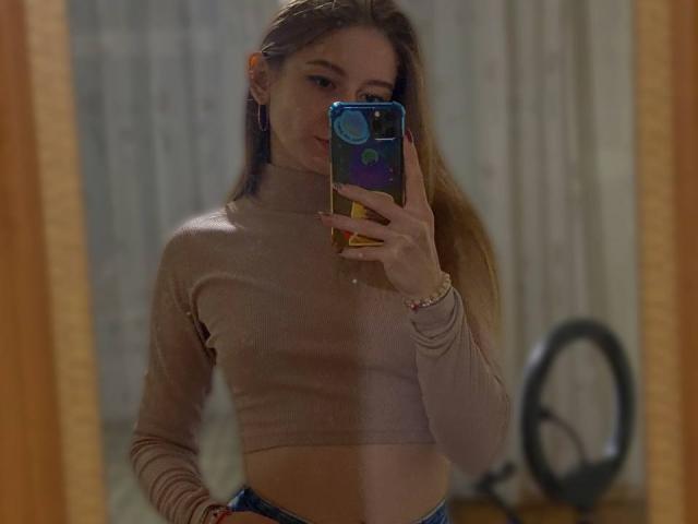 GwynBartell with long blonde hair, wearing a beige turtleneck, taking a mirror selfie with a blue phone, showing her slim waist and small, subtle tits.