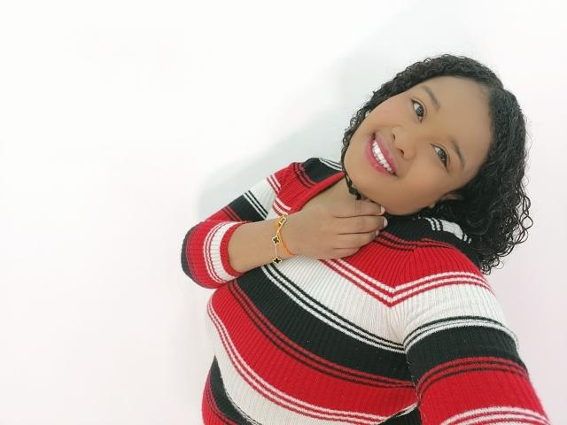Shehyla with curly black hair, smiling, wearing a red, white, and black striped sweater, touching her neck, gold bracelet, white background. Shehyla's tits are not visible.