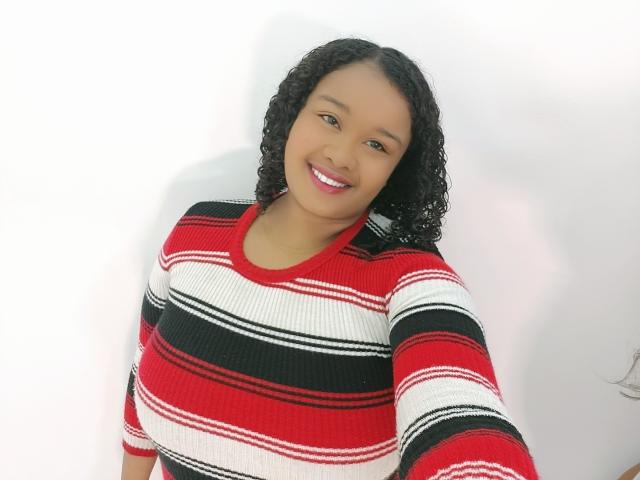 Shehyla, avec ses cheveux noirs bouclés, souriante, portant un pull rayé rouge, blanc et noir, montrant sa poitrine, debout devant un mur blanc, sur une photo de profil..