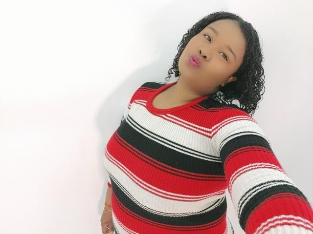 Shehyla, con cabello negro y rizado, vestida con un suéter a rayas rojo, blanco y negro, con labios rosados fruncidos y mirando hacia abajo a la cámara. Sus pechos medianos son parcialmente visibles..