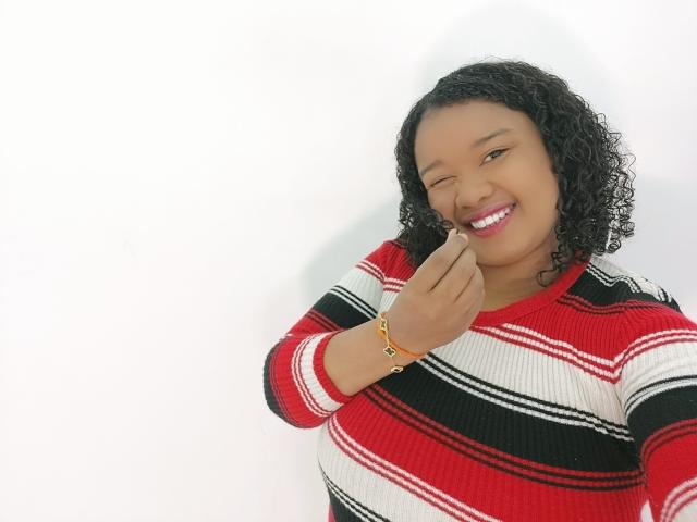 Shehyla, com cabelos pretos encaracolados e batom vermelho, veste um suéter listrado em vermelho, preto e branco, sorrindo e tocando o nariz com o dedo. Ela usa uma pulseira dourada..