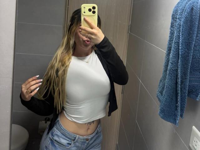 SammyViolet mit langen blonden Haaren, einem weißen T-Shirt, einem schwarzen Cardigan und blauen Jeans, die in einem Spiegel-Selfie im Badezimmer ihre Brüste zeigt, mit hochgezogenen Lippen, und einem gelben iPhone in der Hand..