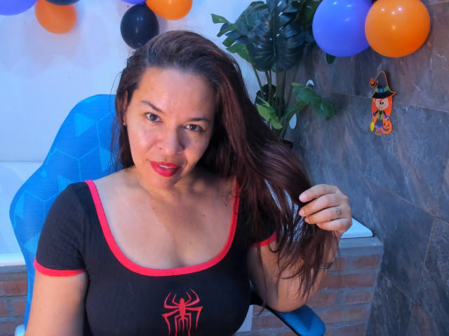 LizethBells with medium brown hair, red lipstick, and black top with red trim and spider emblem, holding hair, sitting in blue chair, surrounded by balloons. Boobs are visible.
