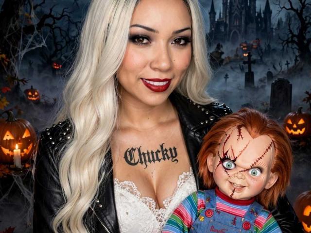 AiunyQueens with platinum blonde hair, red lipstick, black leather jacket, white lace top, showing her tits, holding a creepy Chucky doll with stitched eyes and pumpkin background.