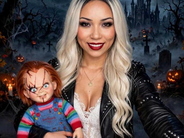 AiunyQueens with long blonde hair, black leather jacket, and cleavage in a white lace top, holding a creepy doll with red hair, blue overalls, and scary face, smiling.