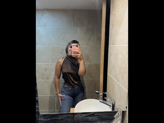 SashaPolet with a slim figure, wearing a sheer black top, blue jeans, and a brown belt, taking a mirror selfie in a bathroom. Her hand is on her hip.