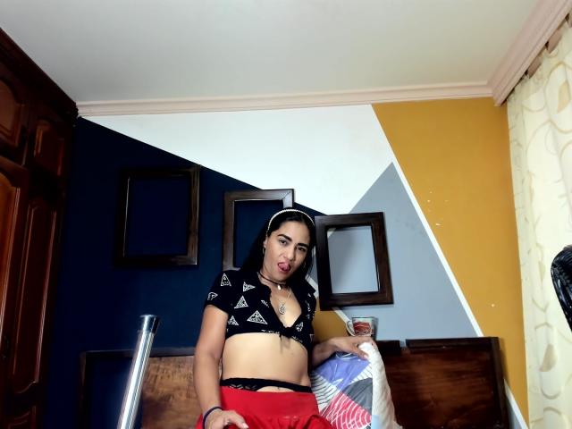 AfroditaHan with medium tits, black triangle crop top, red shorts, holding drink, white headband, yellow wall, dark wood furniture, looking at camera with tongue out.