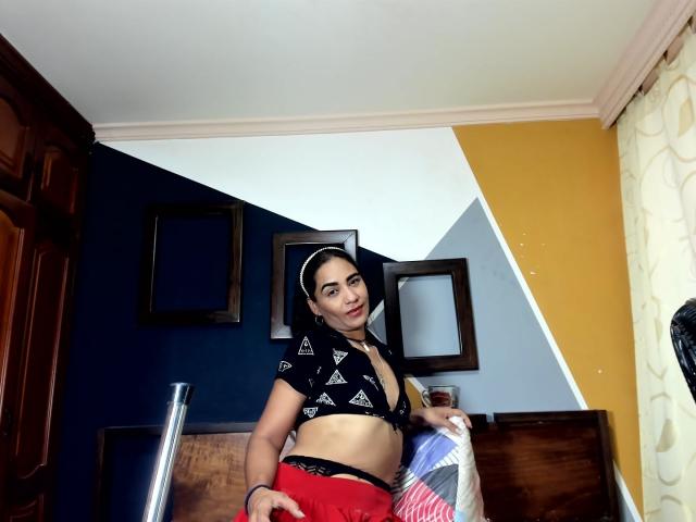 AfroditaHan with small tits, wearing a black triangle pattern crop top and red skirt, standing in a room, looking at the camera, holding a pillow, dark hair, headband.