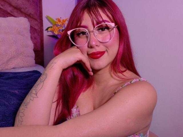 MaddyMills mit rosa Haaren, Brille, rotem Lippenstift und Tattoos, die ihr Kinn in die Hand legt, zeigt ihre Brüste in einem Top mit Blumenmuster und lächelt. Schlafzimmer, violette Beleuchtung..