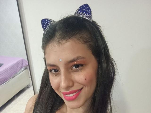 MadissonFontaine with black hair, blue glittery cat ears, pink lipstick, sparkling eyeliner, nose piercing, and a cute smile. Boobs not visible. Bed with purple cover in background.